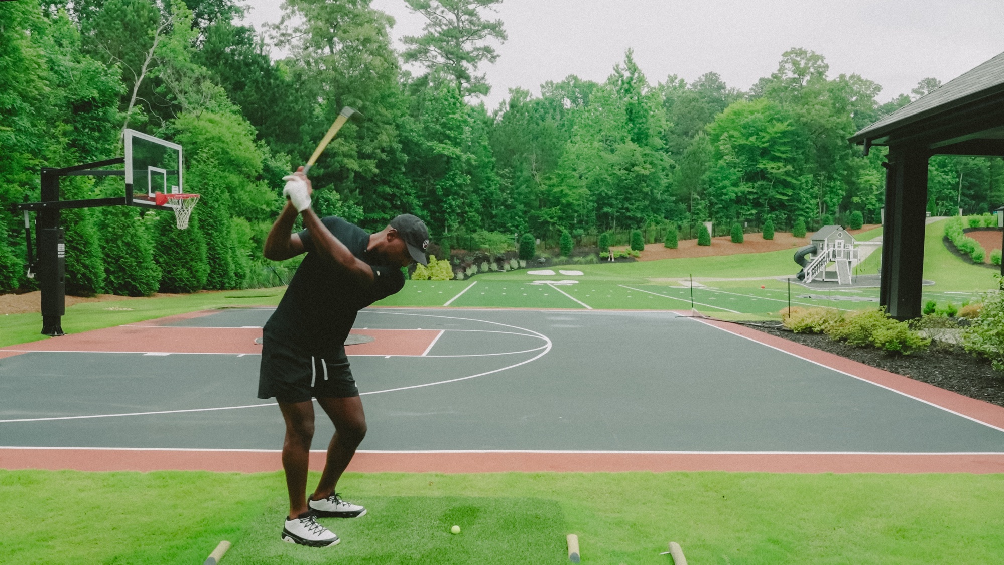 Patrick Peterson at the top of his backswing on a backyard tee beside a basketball court, hitting toward a full-size replica golf hole
