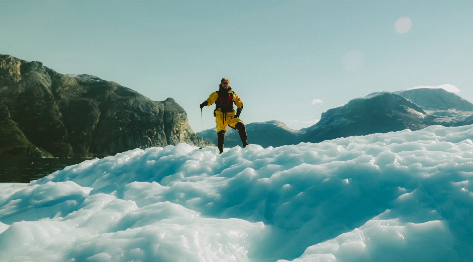 A golfer in a yellow drysuit stands atop an iceberg with a club in hand, mountains of a Greenland fjord behind him